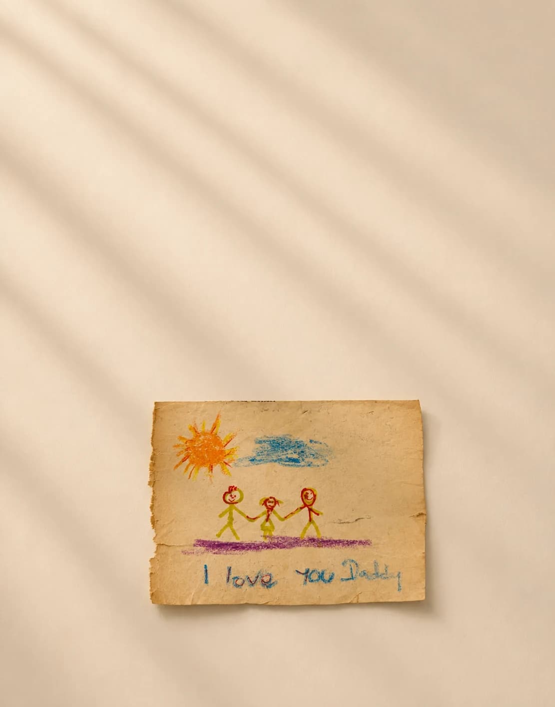 Book-style image of a crayon drawing on worn paper with a family under the sun and the words I love you Daddy.